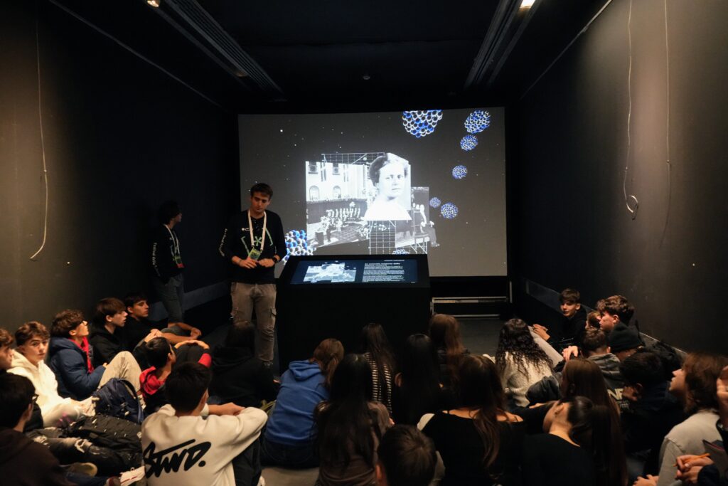 ragazzi seduti per terra che asoltano animatore mostra Viaggio alm centro dell'atomo con immagini atomo e Lise Meitner sullo schermo
