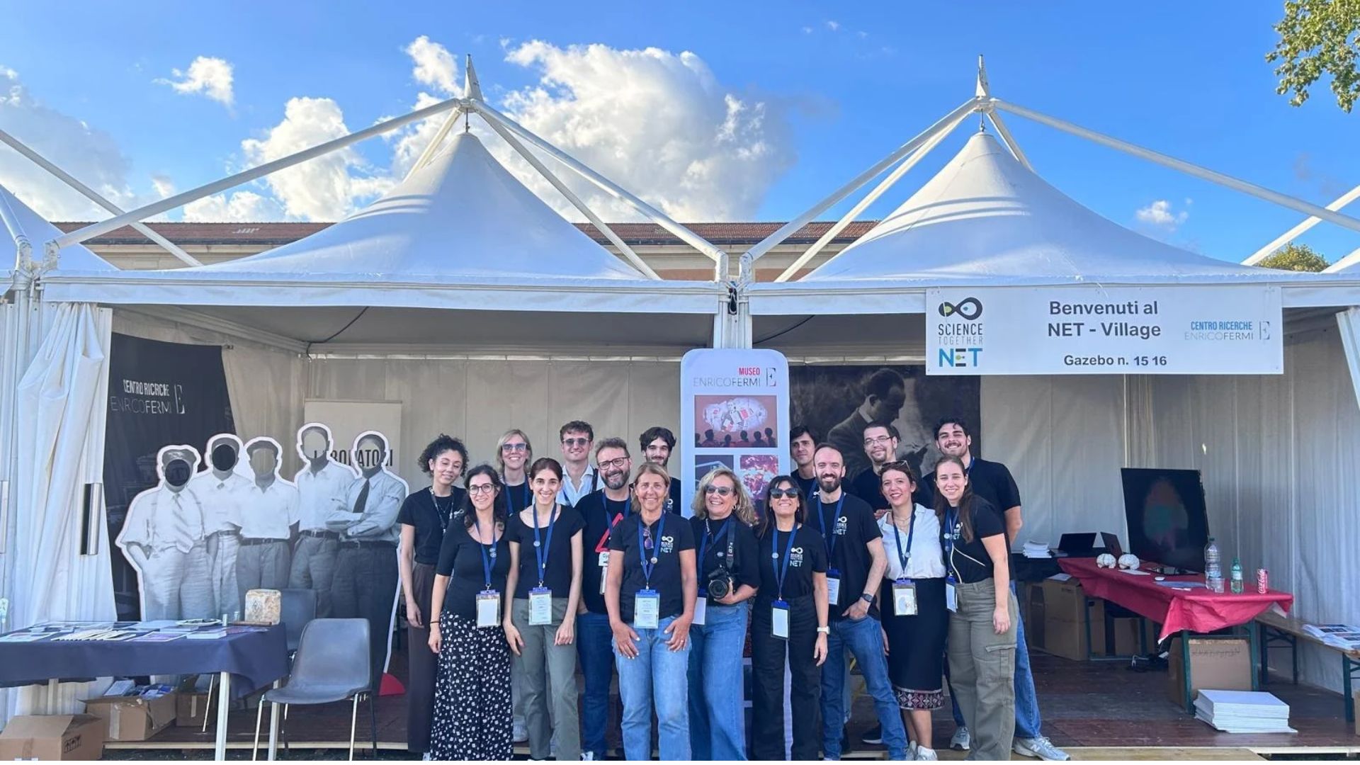 Staff del Centro Ricerche Enrico Fermi in maglia nera davanti ai gazebo 15-16 del NET Village, con cartello "Benvenuti al NET - Village