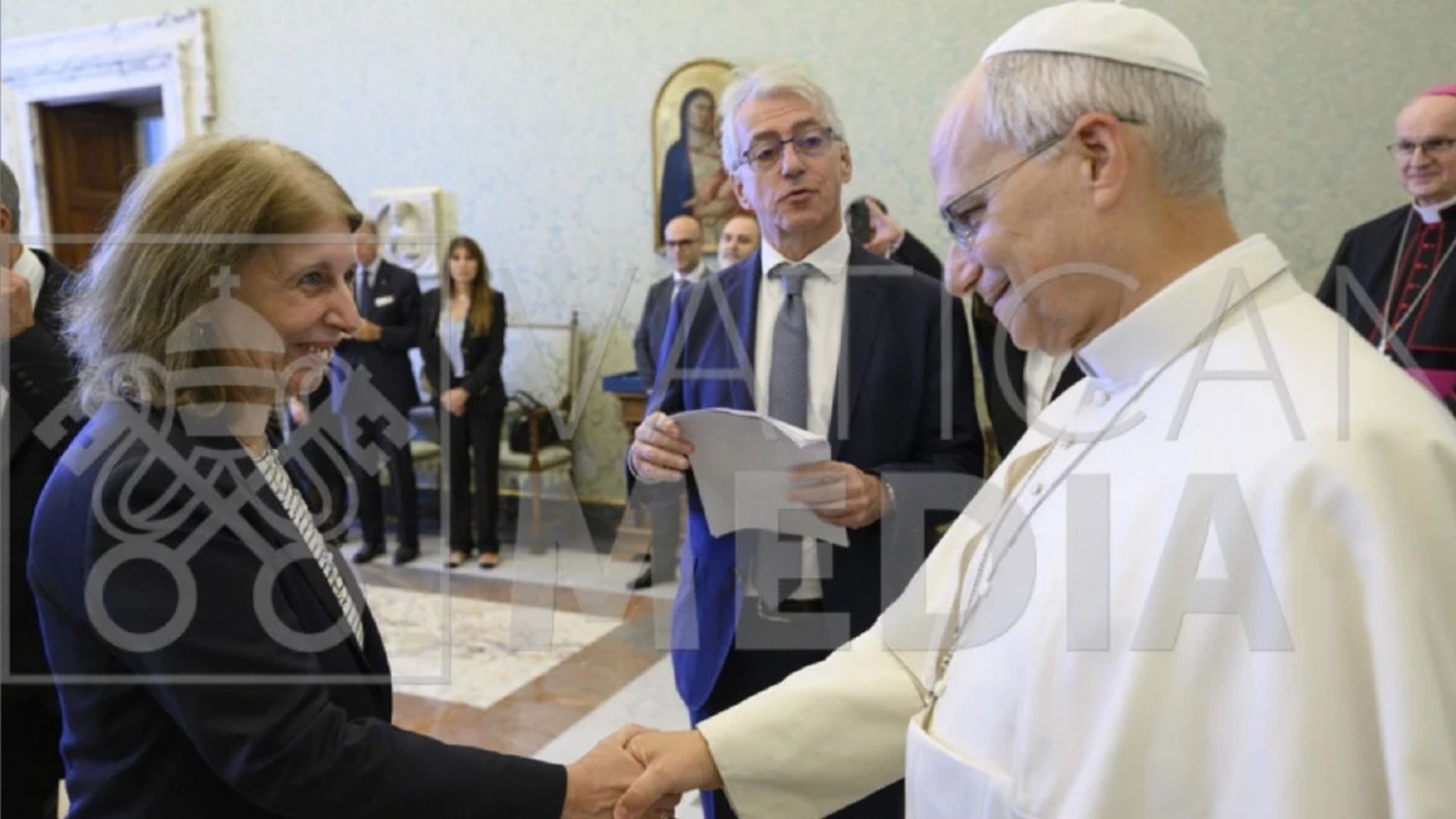 Il Papa stringe la mano a una donna sorridente durante un'udienza in Vaticano; accanto, un uomo con dei fogli in mano