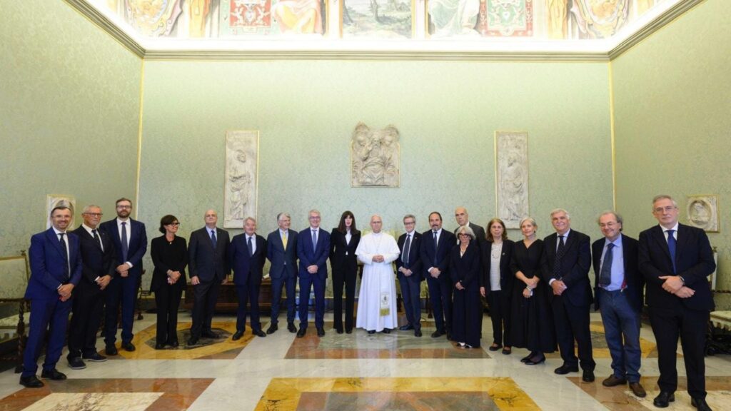 Il Papa in abito bianco posa con un gruppo di persone in abiti scuri in una sala con affreschi e bassorilievi